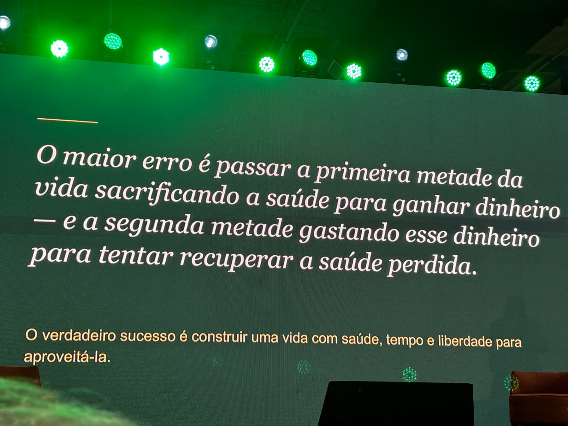 Telão de palestra com a frase: “O maior erro é passar a primeira metade da vida sacrificando a saúde para ganhar dinheiro — e a segunda metade gastando esse dinheiro para tentar recuperar a saúde perdida.”